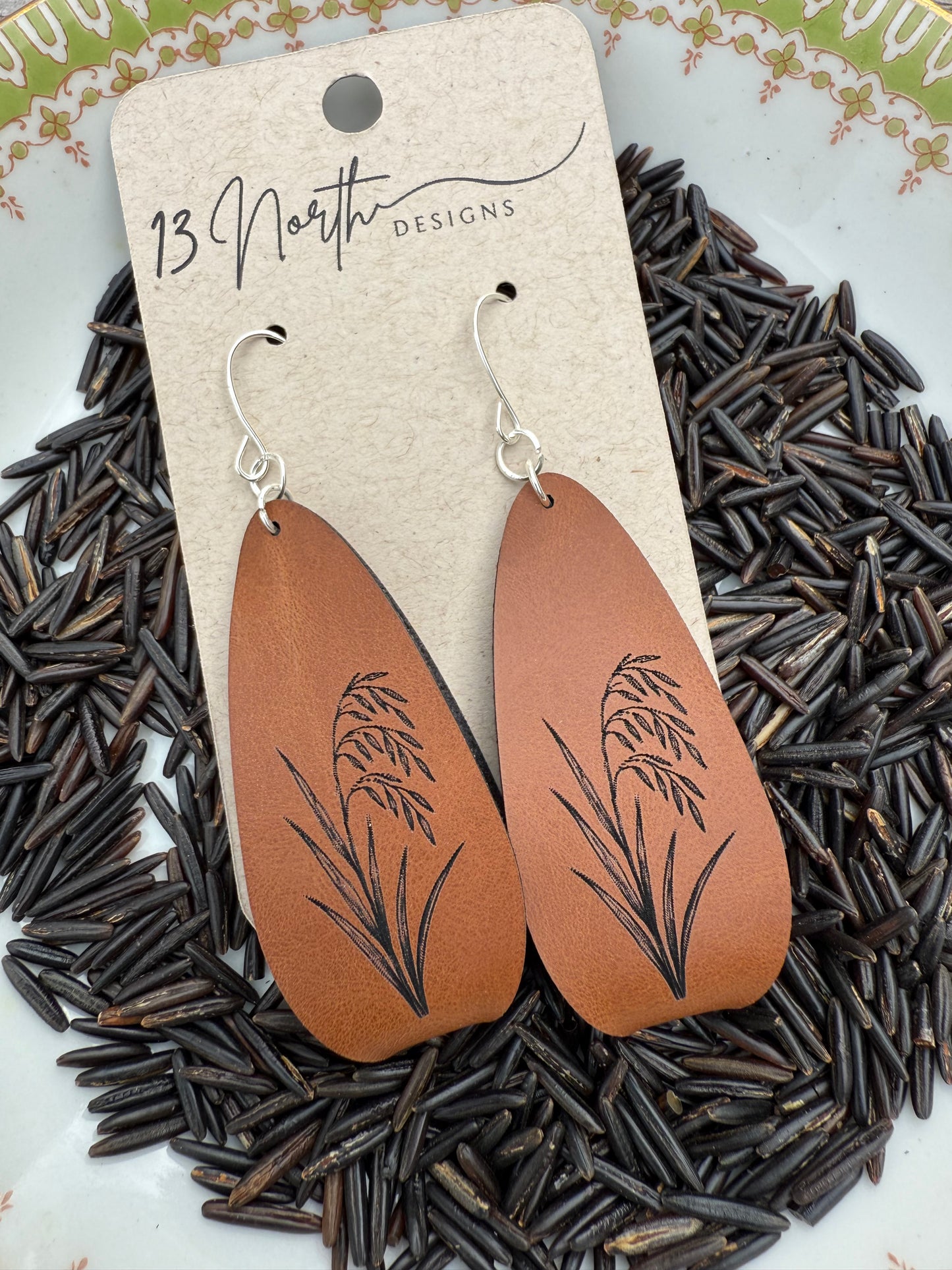 Brown leather earrings with engraved wild rice on a card, placed on a bed of wild rice