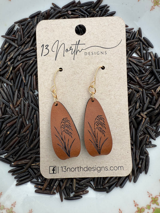 Brown leather earrings with wild rice plant on a beige card, placed on a bed of wild rice.